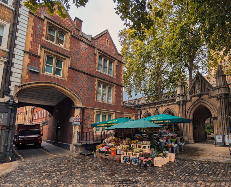 An outdoor flower stall situated on a cobblestone street in front of a historic brick and stone building with Gothic architectural details, featuring an arched entrance and decorative stonework. The s