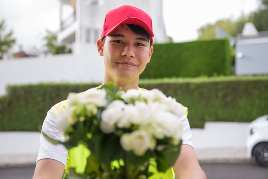 A delivery person wearing a red cap and a high-visibility vest is smiling while holding a bouquet of fresh white and green flowers, including roses and lilies, wrapped in clear cellophane. The bouquet